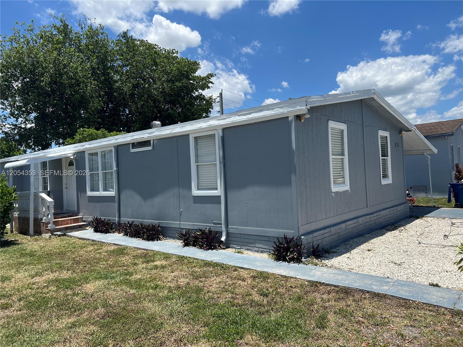 A gray mobile home features white trim, a gravel driveway, and a small front porch — 20016 NW 56 Ave, Miami Gardens FL 33055, MIAMI GARDENS, FL