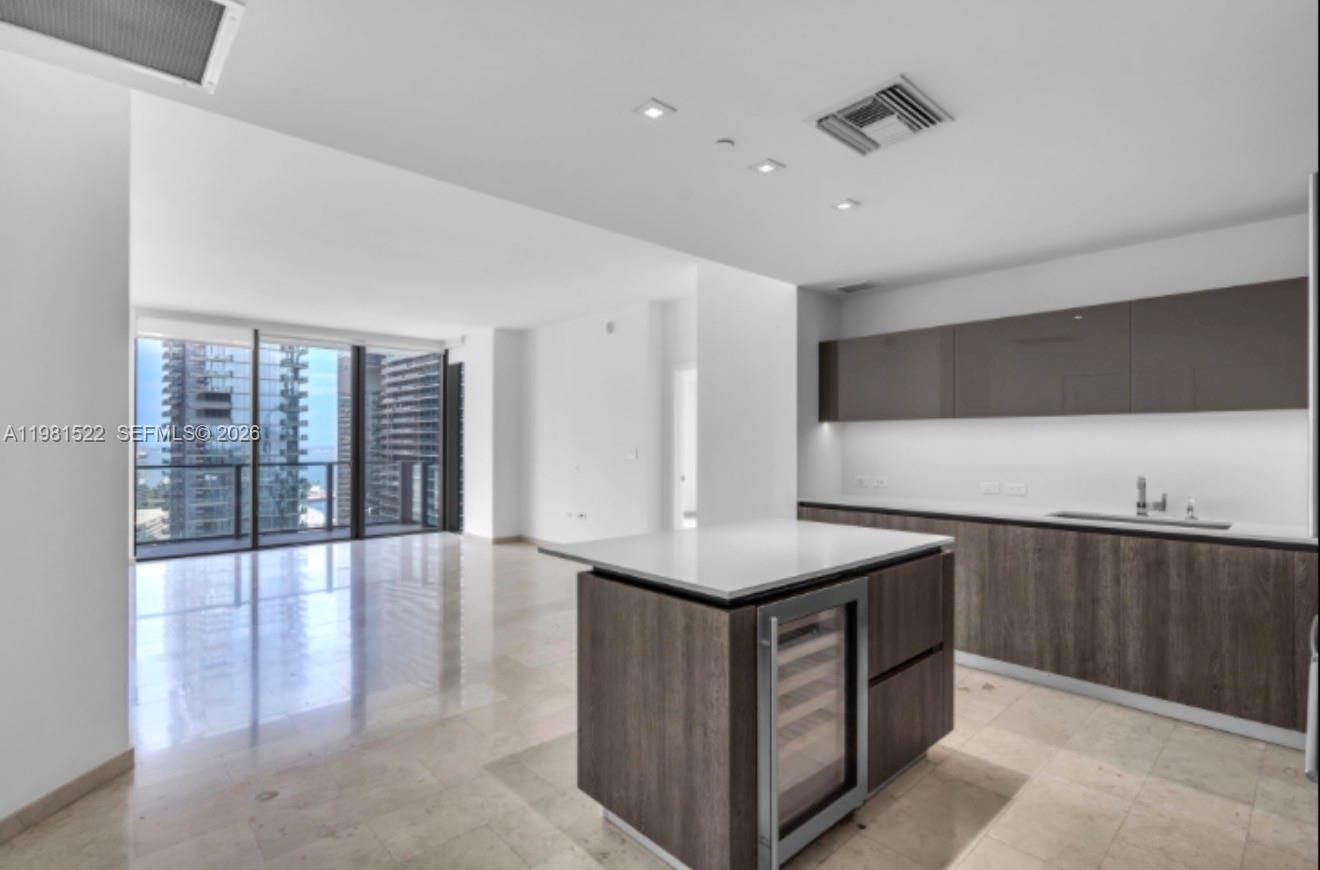 A modern kitchen with wood-toned cabinetry and a central island overlooks a bright living area — 88 SW 7TH ST # 2005, Miami FL 33130, MIAMI, FL