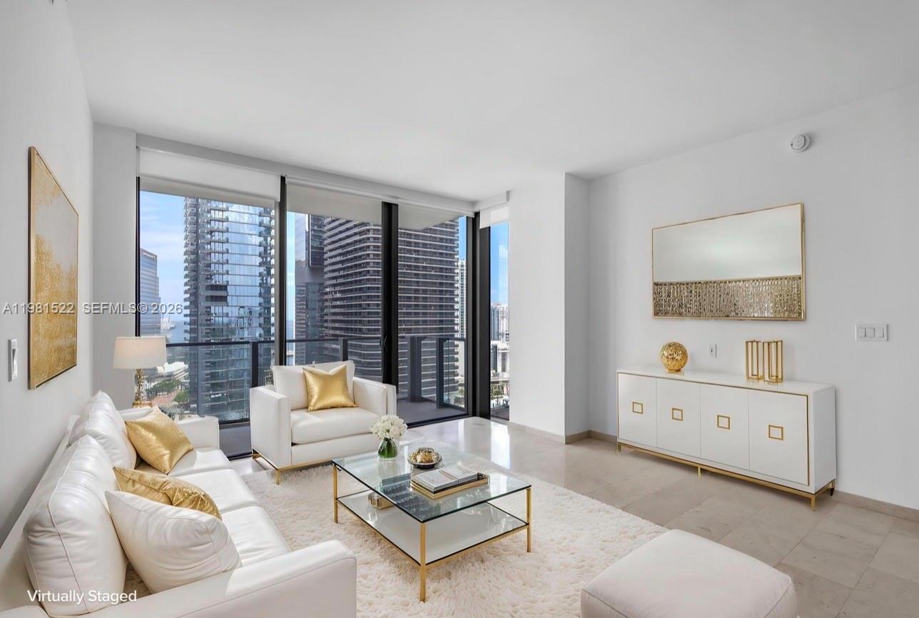 A modern living room features white furniture, gold accents, and floor-to-ceiling windows with city views — 88 SW 7TH ST # 2005, Miami FL 33130, MIAMI, FL