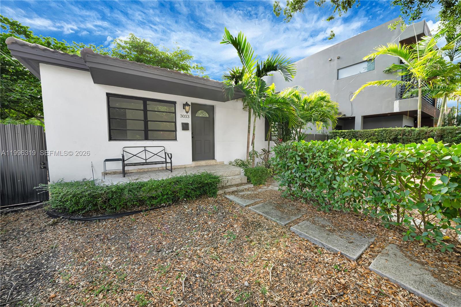 A white single-story house features a dark front door, a metal bench, and landscaping — 3033 Hibiscus St, Miami FL 33133, MIAMI, FL