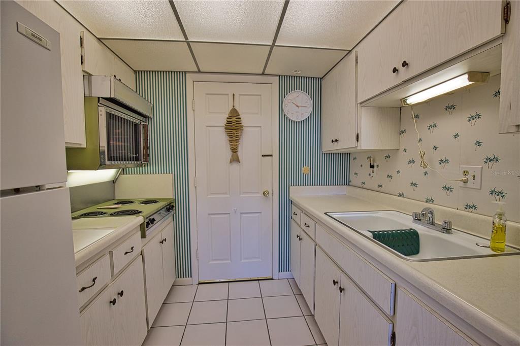 A galley kitchen features white cabinets, striped wallpaper, a white sink, and tiled flooring — 200 N THE ESPLANADE UNIT C-9, VENICE, FL