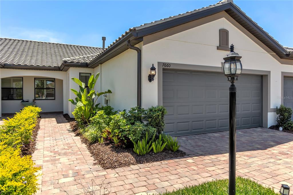 A single-story house exterior features a gray garage door, brick paver driveway, and landscaping — 7660 SUMMERLAND COVE, BRADENTON, FL