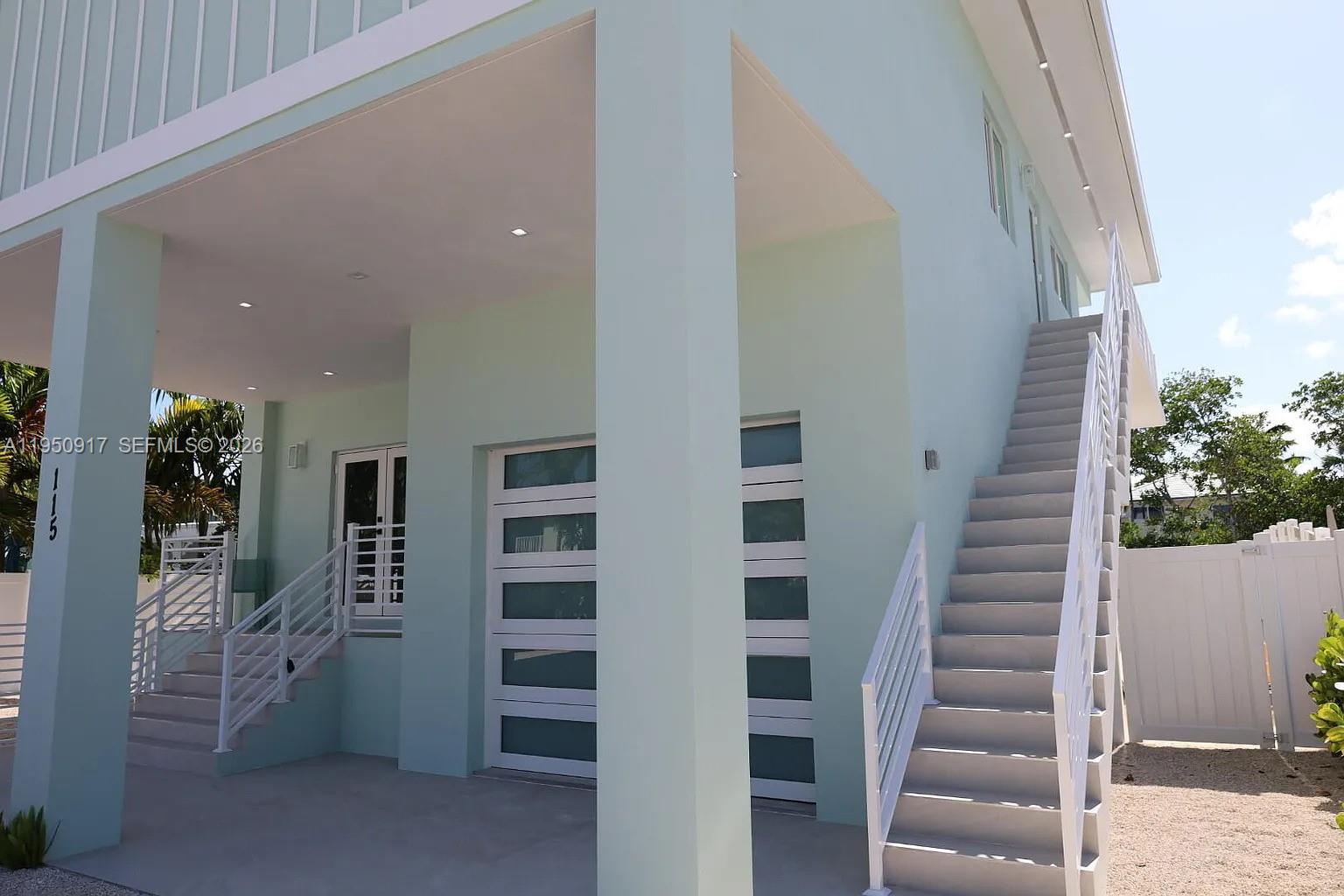 A light green elevated house features an exterior staircase, a covered patio, and garage doors — 115 Azalea St, Plantation Key FL 33070, PLANTATION KEY, FL