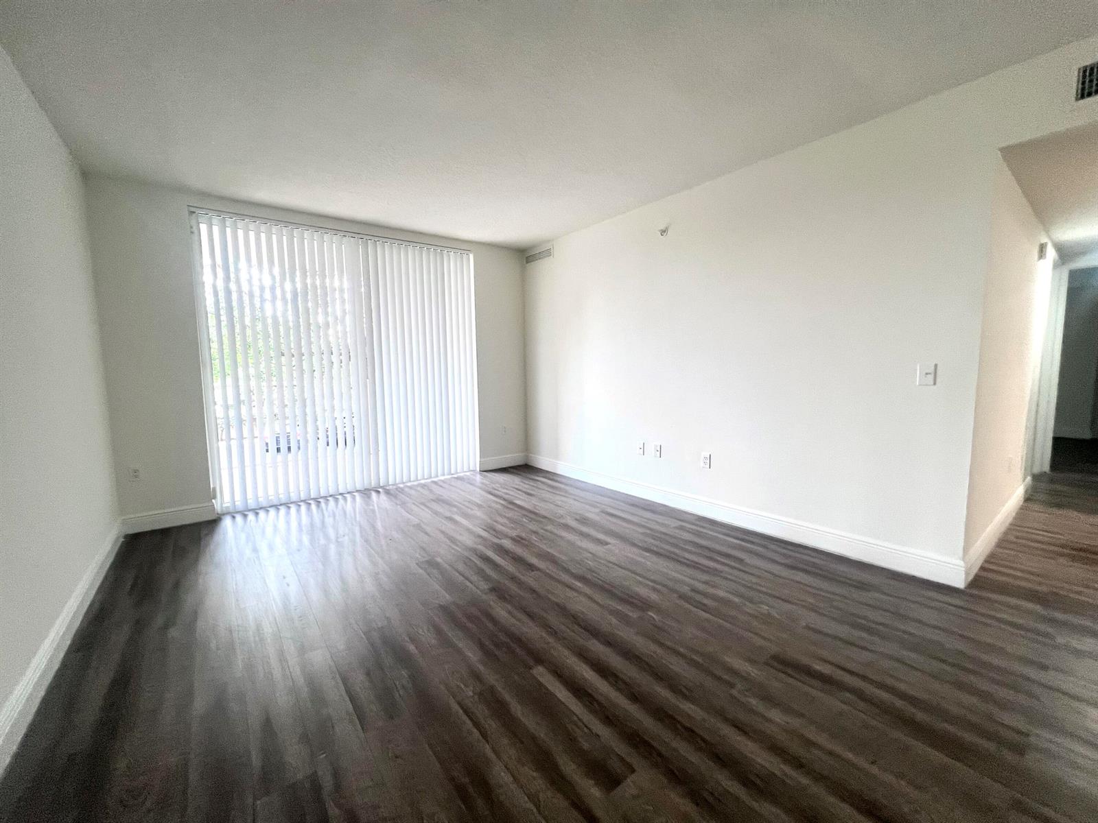 An empty living room features dark wood-look flooring, white walls, and vertical blinds over a door — 18335 NW 68 Avenue # 110, Hialeah FL 33015, HIALEAH, FL