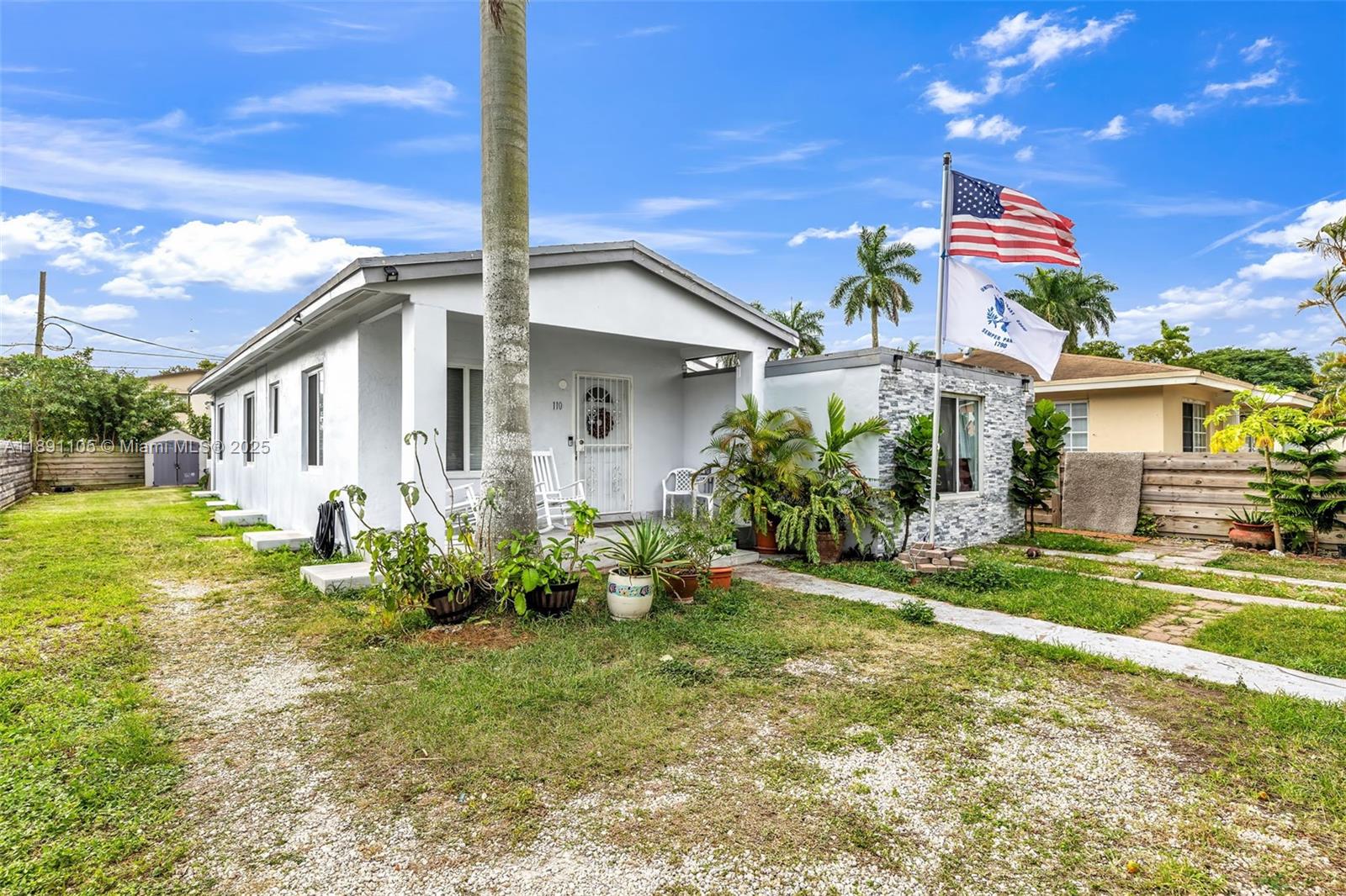 A white single-story house features a front porch, a gravel yard, and two flying flags — 110 NE 9th Ct, Homestead FL 33030, HOMESTEAD, FL