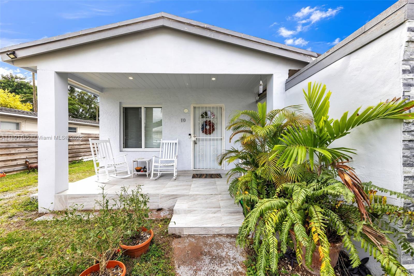 A white house exterior features a covered porch with two rocking chairs and a palm plant — 110 NE 9th Ct, Homestead FL 33030, HOMESTEAD, FL