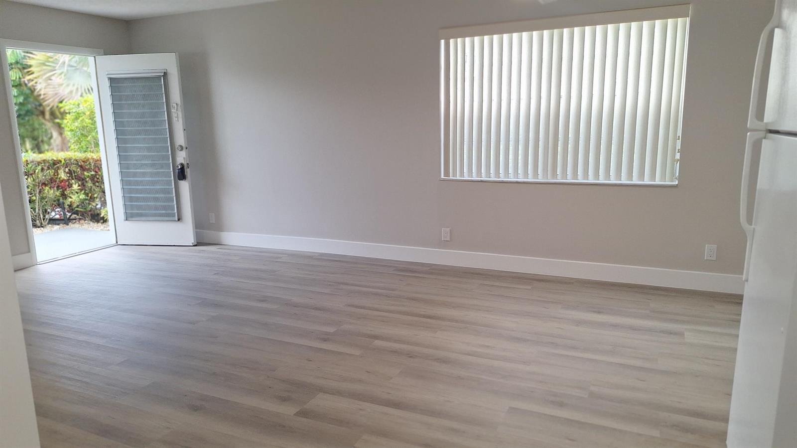 An empty living room features light wood-look flooring, gray walls, vertical blinds, and an open door — 120 Normandy # C, Delray Beach FL 33484, DELRAY BEACH, FL