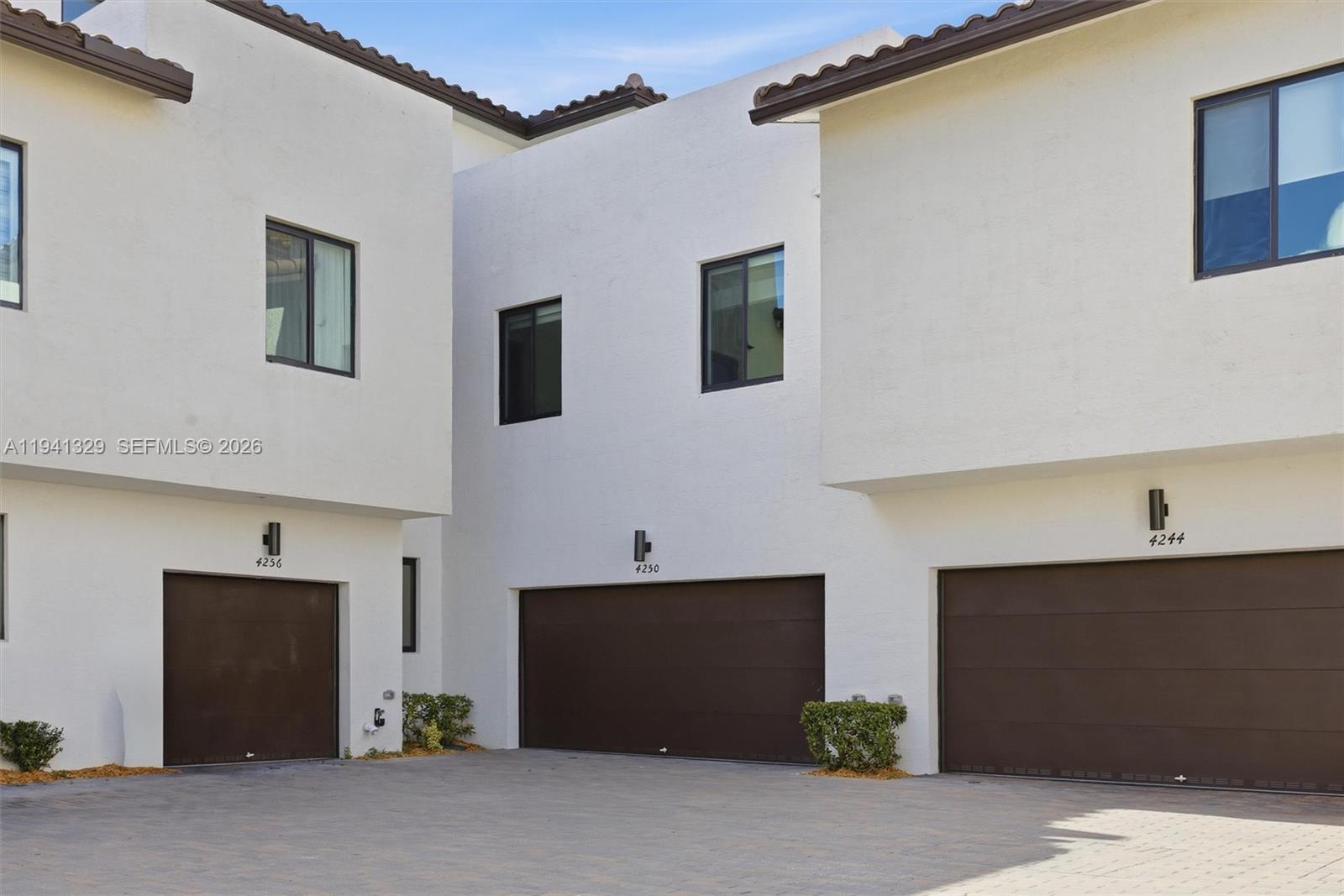 Three white townhouses with brown garage doors and dark-framed windows stand on a paved lot — 4250 NW 83rd Ave, Doral FL 33166, DORAL, FL