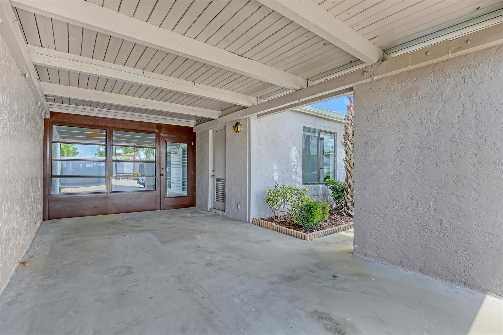 A covered carport features a concrete floor, a white wood-beam ceiling, and a stucco wall — 14735 Wildflower Lane, Delray Beach FL 33446, DELRAY BEACH, FL