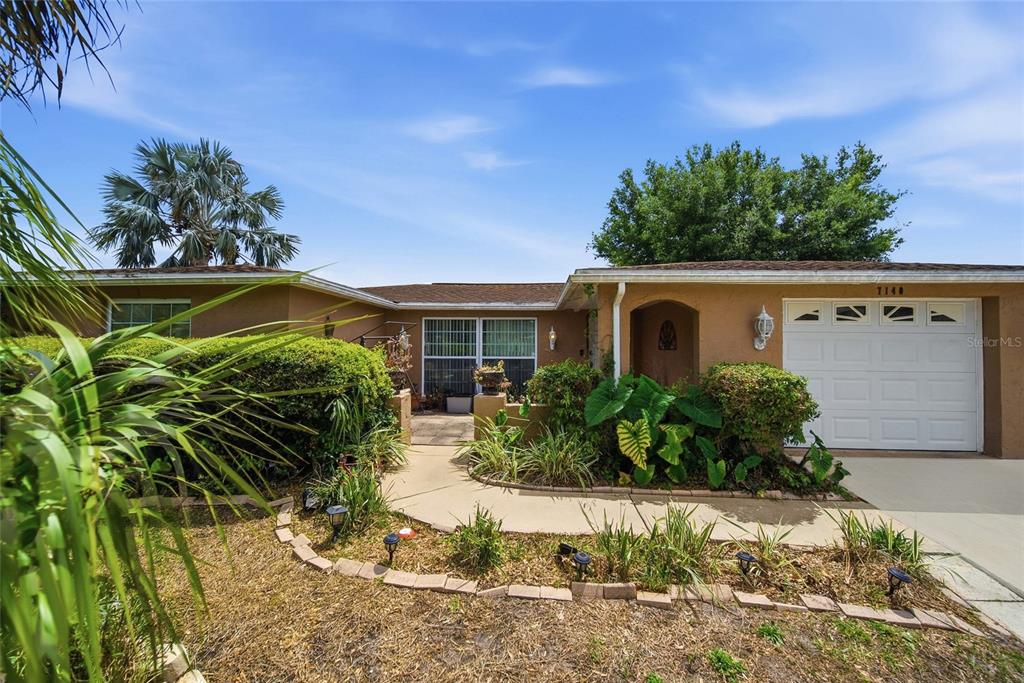 A single-story house with a tan stucco exterior, a white garage door, and landscaped front yard — 7140 ROCKWOOD DRIVE, PORT RICHEY, FL