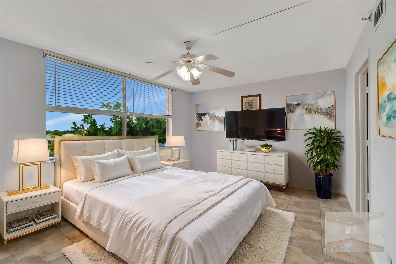 A bedroom features a white bed, a dresser with a television, and large windows — 14623 Bonaire Boulevard # 502, Delray Beach FL 33446, DELRAY BEACH, FL