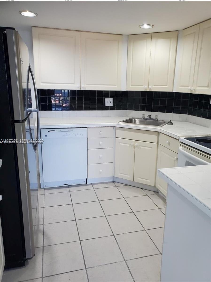 A kitchen features cream cabinets, a black tiled backsplash, white countertops, and white tile flooring — 3301 Spanish Moss Ter # 708, Lauderhill FL 33319, LAUDERHILL, FL