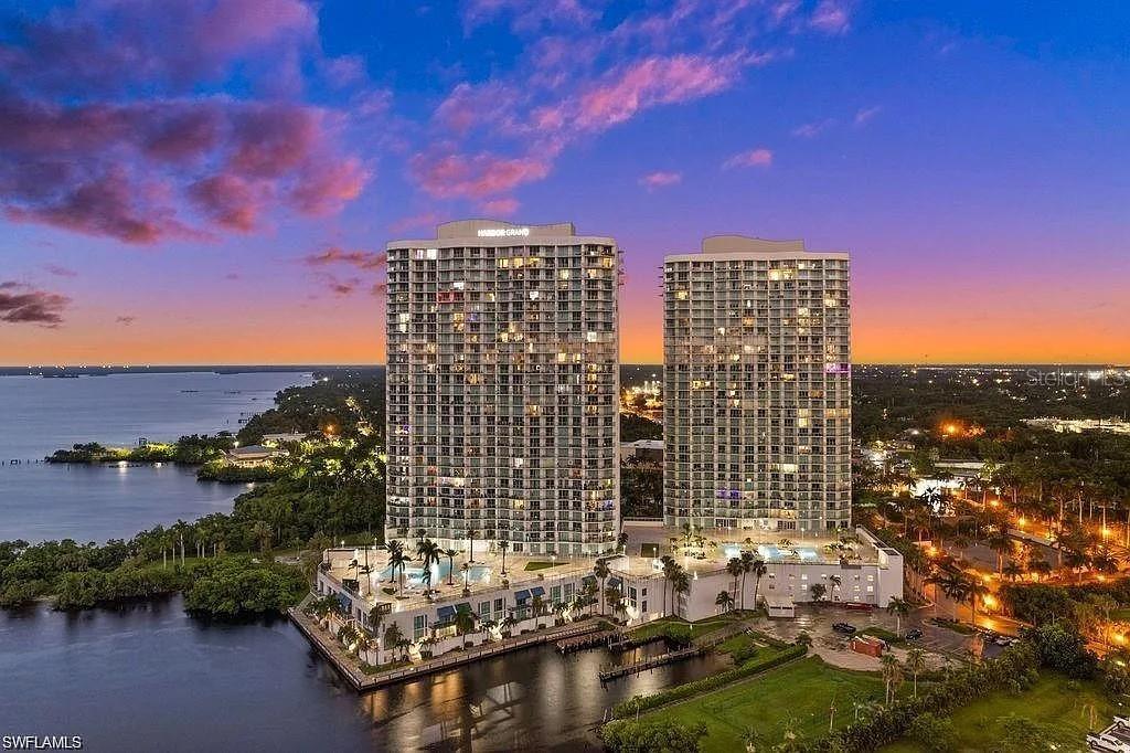 An aerial view shows two tall residential towers situated along a waterfront at sunset — 3000 OASIS GRAND BOULEVARD UNIT 1002, FORT MYERS, FL