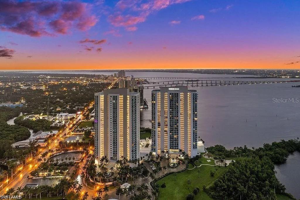 An aerial view shows two tall residential towers situated along a waterfront at sunset — 3000 OASIS GRAND BOULEVARD UNIT 1002, FORT MYERS, FL