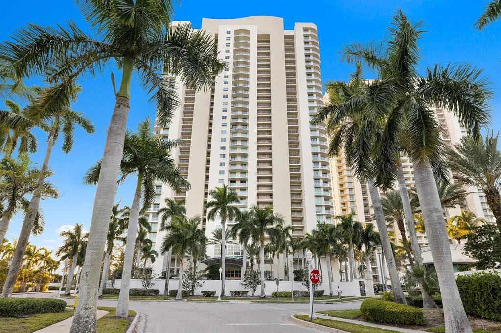 A tall, beige residential high-rise building surrounded by palm trees under a clear blue sky — 3000 OASIS GRAND BOULEVARD UNIT 1002, FORT MYERS, FL