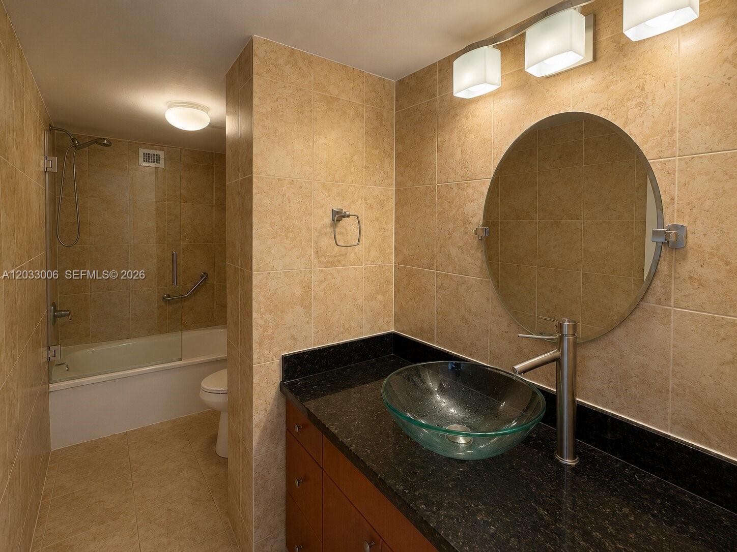 A bathroom features a glass vessel sink, black countertop, round mirror, and a tiled tub — 1000 Parkview Dr # 415, Hallandale Beach FL 33009, HALLANDALE BEACH, FL