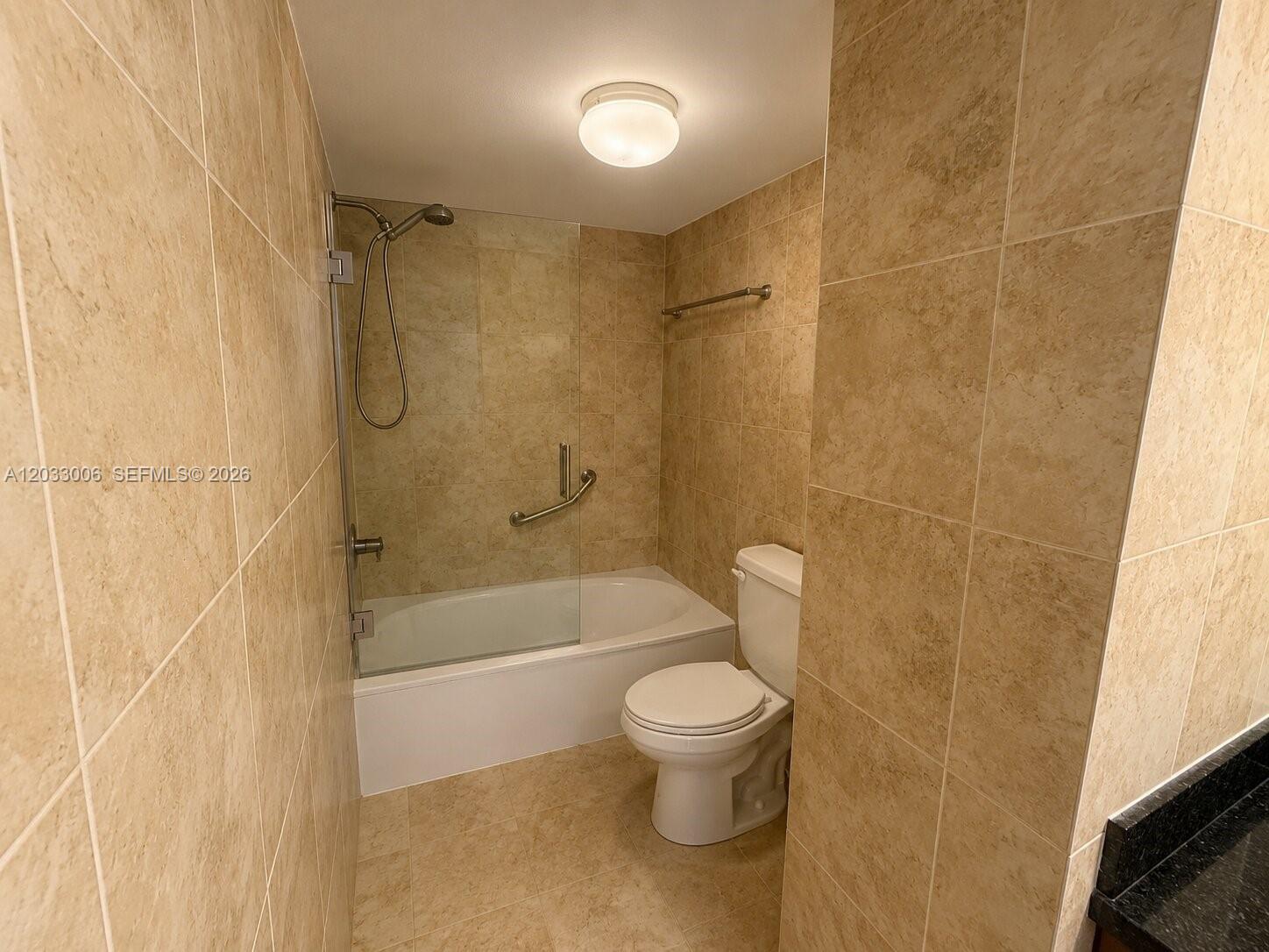 A bathroom features beige tiled walls, a glass-enclosed shower-tub combination, and a white toilet — 1000 Parkview Dr # 415, Hallandale Beach FL 33009, HALLANDALE BEACH, FL