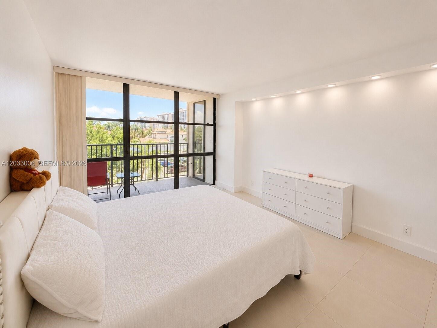 A bedroom features a white bed, a matching dresser, and a balcony with outdoor seating — 1000 Parkview Dr # 415, Hallandale Beach FL 33009, HALLANDALE BEACH, FL