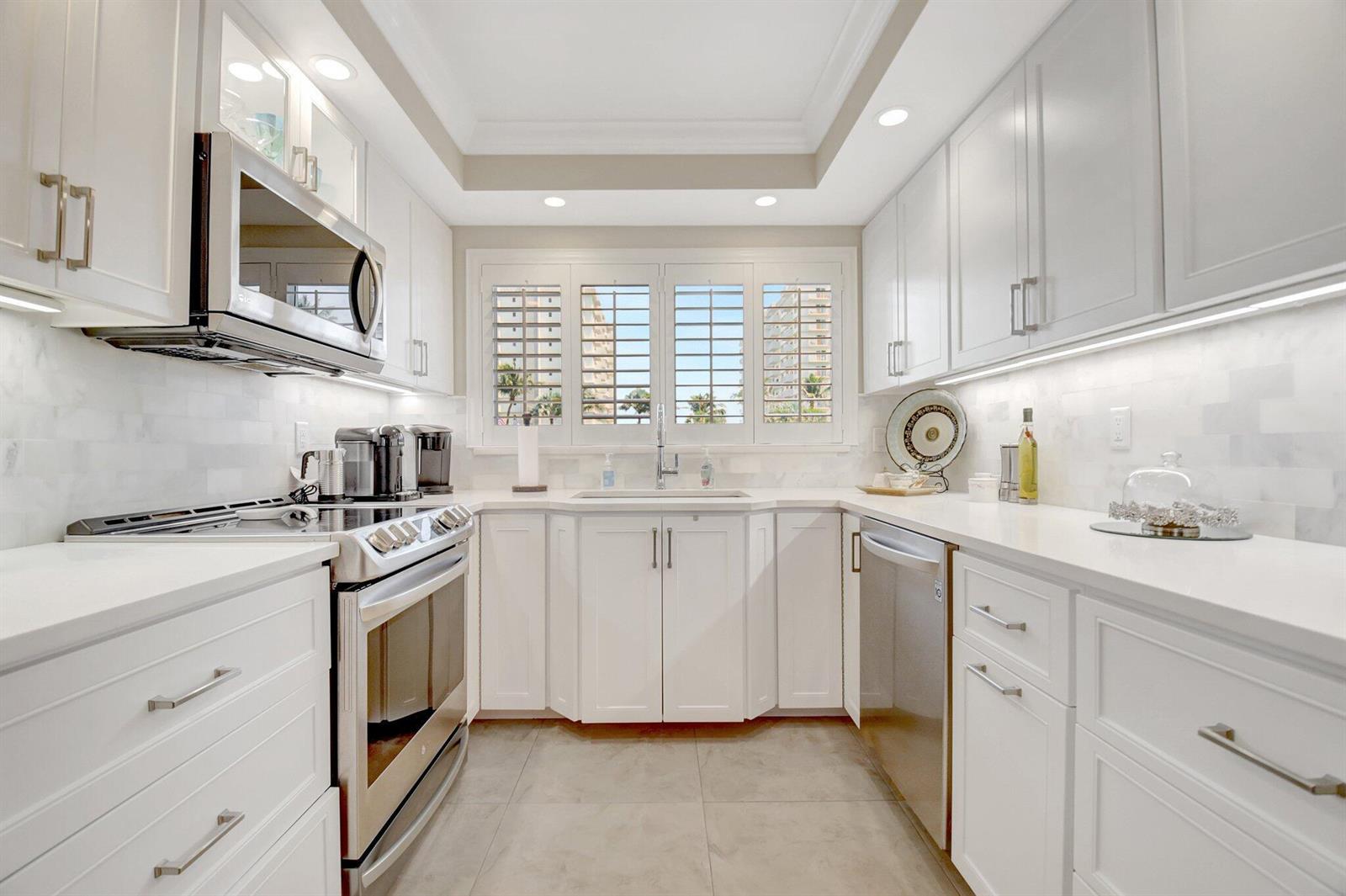 A modern white kitchen features stainless steel appliances, quartz countertops, and plantation shutters over the sink — 3224 S Ocean Boulevard # 214, Highland Beach FL 33487, HIGHLAND BEACH, FL