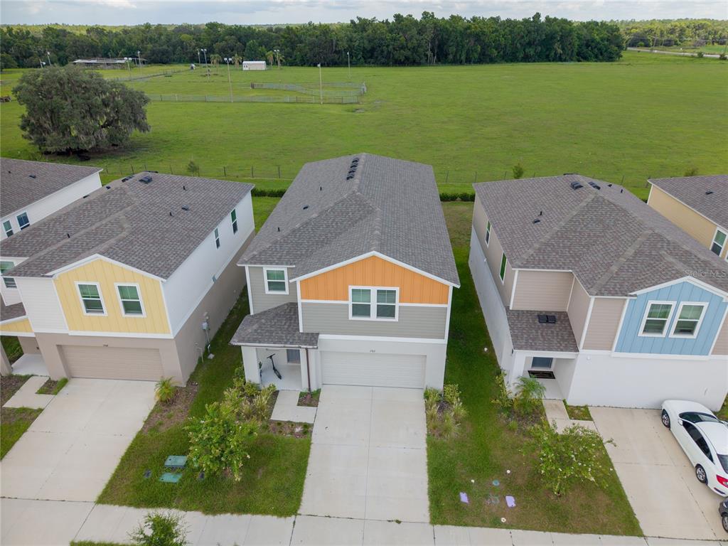 An aerial view shows three suburban houses with driveways facing a large, open green field — 7917 PEACE LILY AVENUE, WESLEY CHAPEL, FL