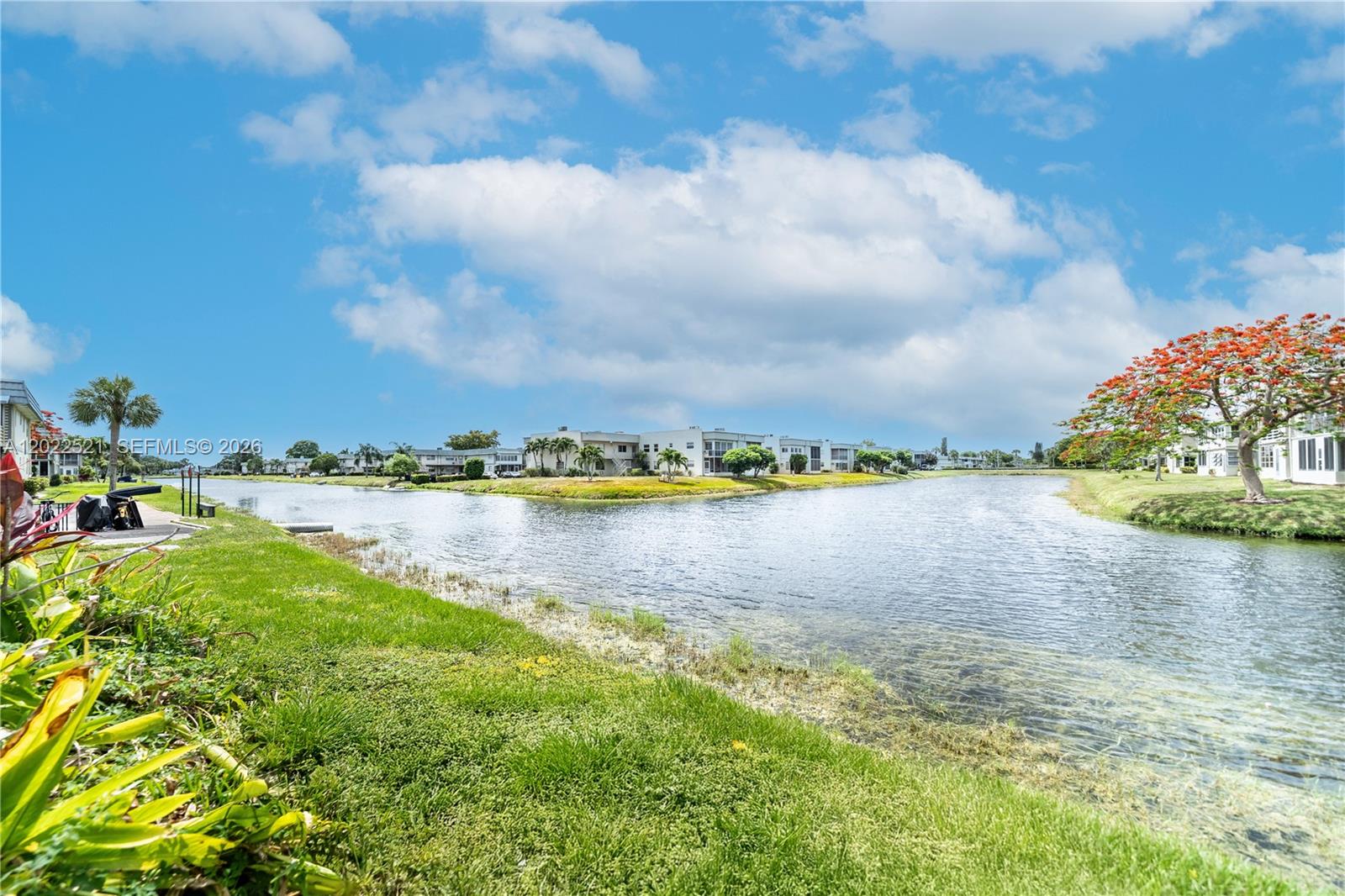 A canal flows between grassy banks with palm trees and residential buildings under a cloudy sky — 145 Brittany D # 145, Delray Beach FL 33446, DELRAY BEACH, FL