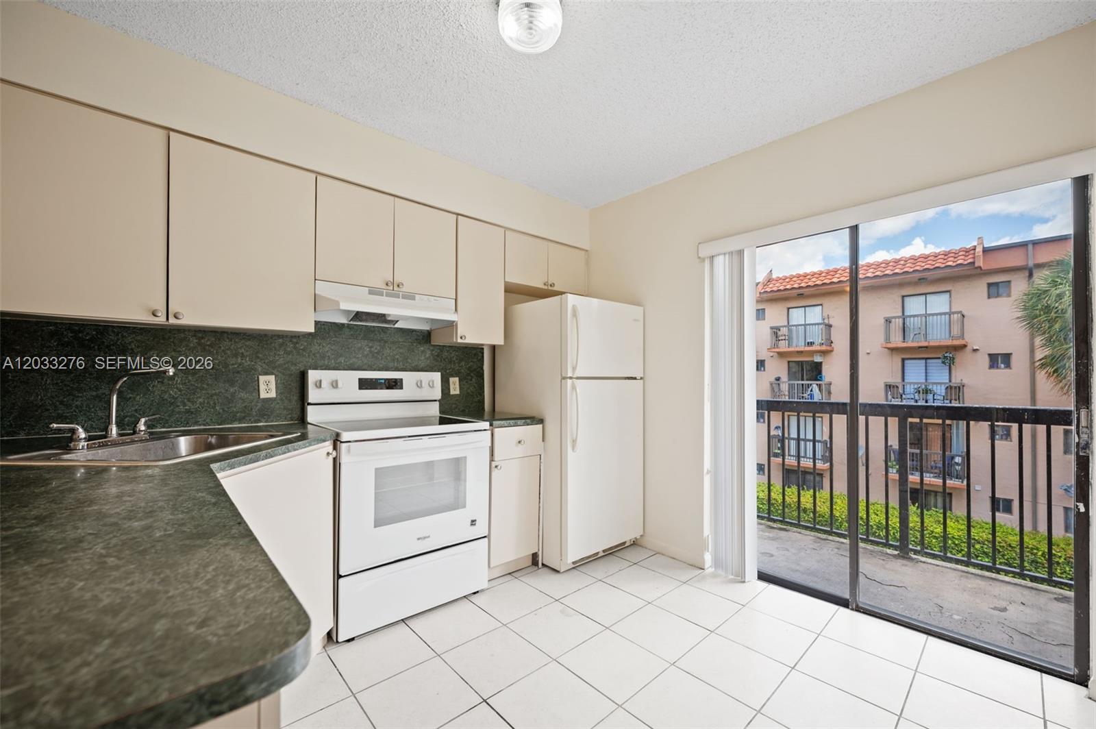 A kitchen with white appliances, beige cabinets, dark countertops, and a sliding glass balcony door — 5490 W 21st Ct # 310, Hialeah FL 33016, HIALEAH, FL