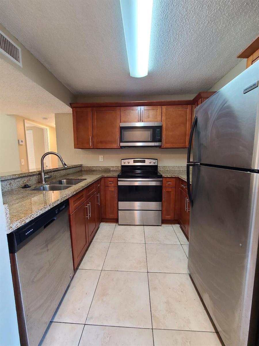 A galley kitchen features stainless steel appliances, wood cabinets, and granite countertops — 1461 Windorah Way # G, West Palm Beach FL 33411, WEST PALM BEACH, FL