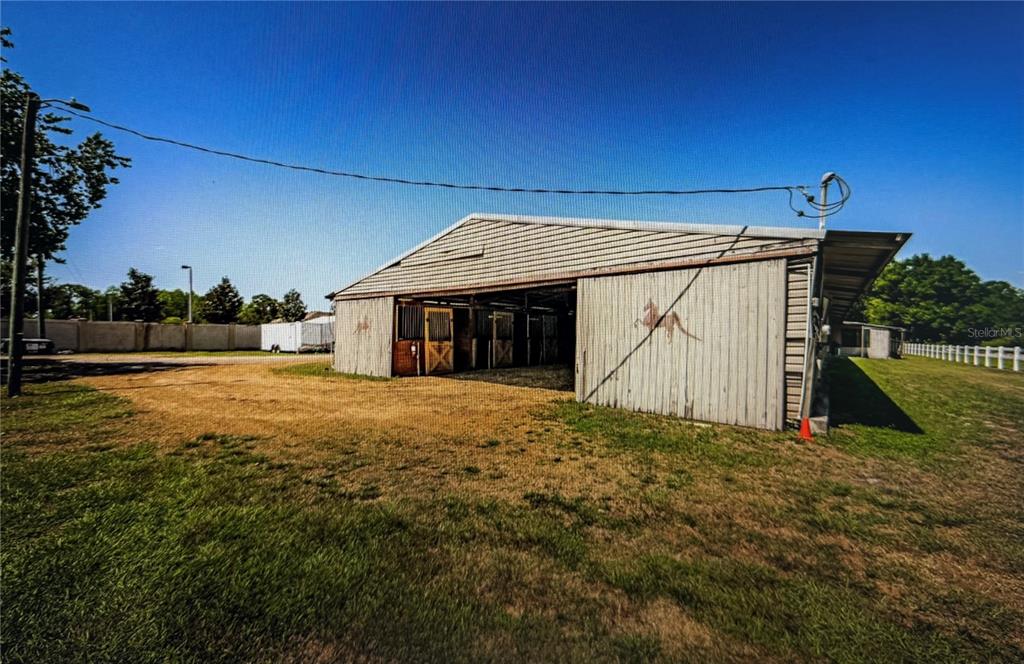 A metal-sided horse barn with open stalls stands on a grassy lot with fencing — 10502 HENDERSON ROAD, TAMPA, FL