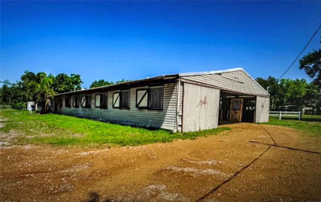 A long, white-sided horse barn with multiple stall windows sits on a gravel lot — 10502 HENDERSON ROAD, TAMPA, FL