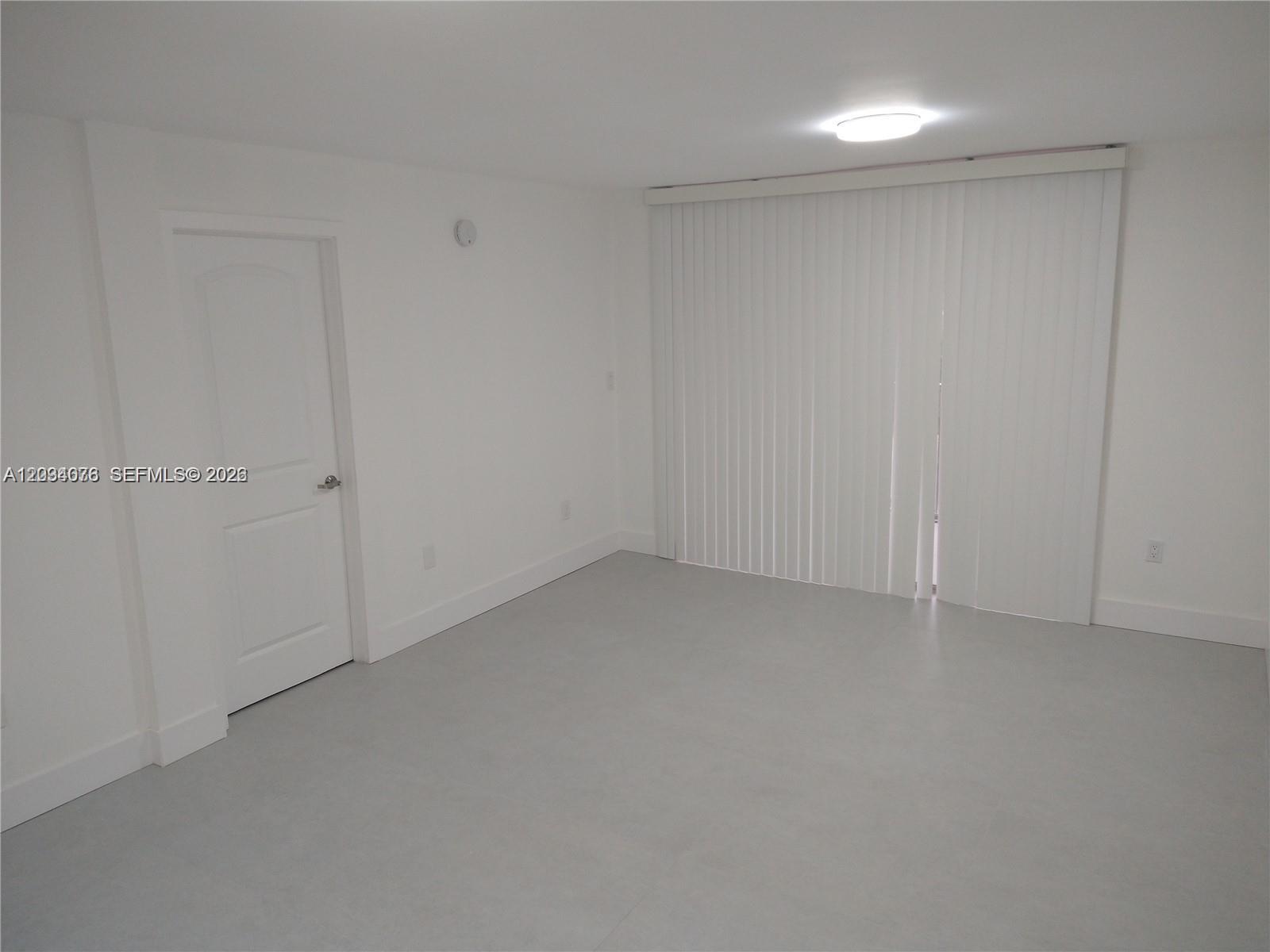 An empty room with white walls, gray flooring, a closed door, and vertical window blinds — 19791 SW 114th Ave # 233, Miami FL 33157, MIAMI, FL