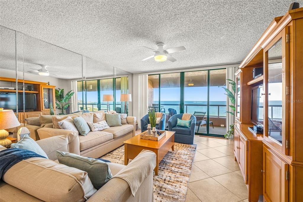 A living room features a beige sofa, blue armchairs, mirrored walls, and a balcony overlooking the ocean — 1310 GULF BOULEVARD UNIT 10D, CLEARWATER BEACH, FL