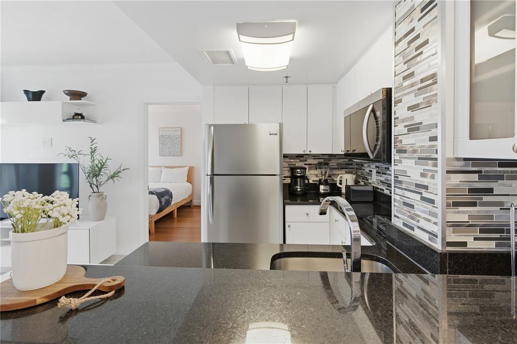 A kitchen with white cabinets, stainless steel appliances, and black granite countertops overlooks a bedroom — 6165 CARRIER DRIVE UNIT 1304, ORLANDO, FL