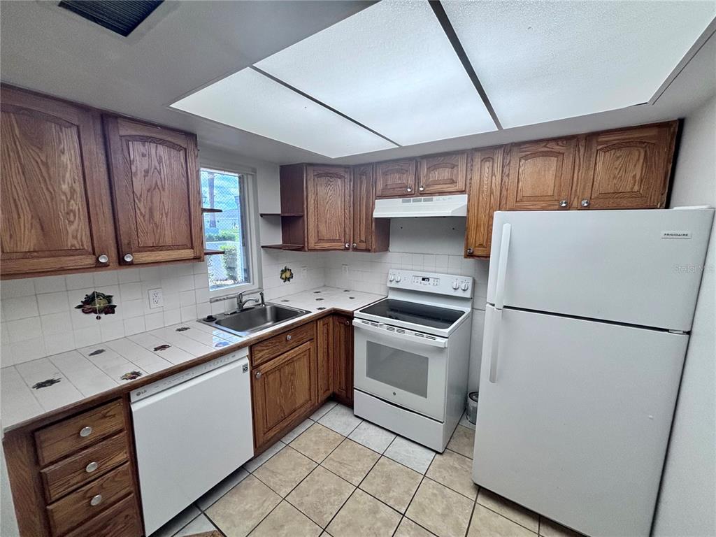 A kitchen features oak cabinets, white appliances, a tiled countertop, and a drop ceiling — 576 FAIRWAYS LANE UNIT M104, OCALA, FL
