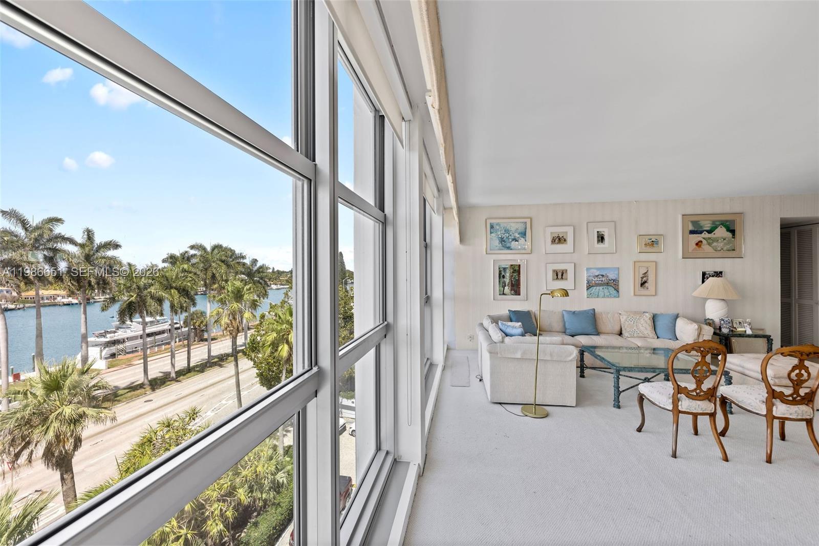 A living room with white carpet and wall art overlooks a canal and palm trees — 5055 Collins Ave # 5M, Miami Beach FL 33140, MIAMI BEACH, FL