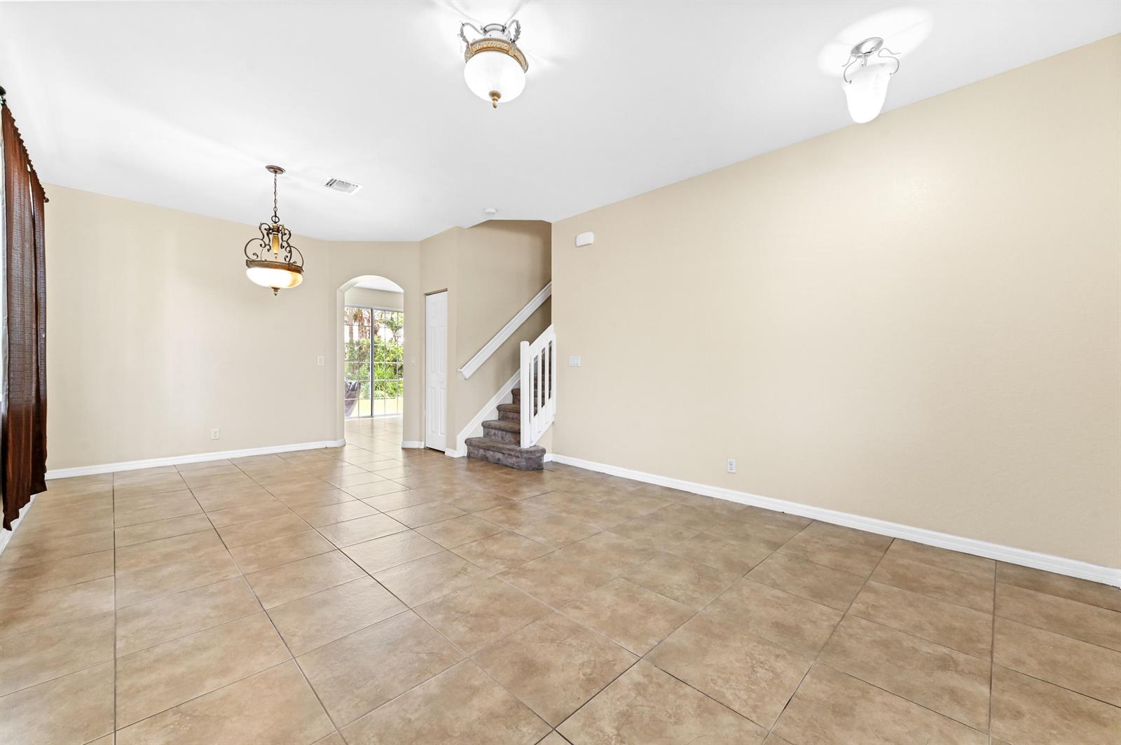 A spacious, empty room with beige walls, tiled floors, a staircase, and two hanging light fixtures — 6819 Osage Circle, West Palm Beach FL 33413, WEST PALM BEACH, FL