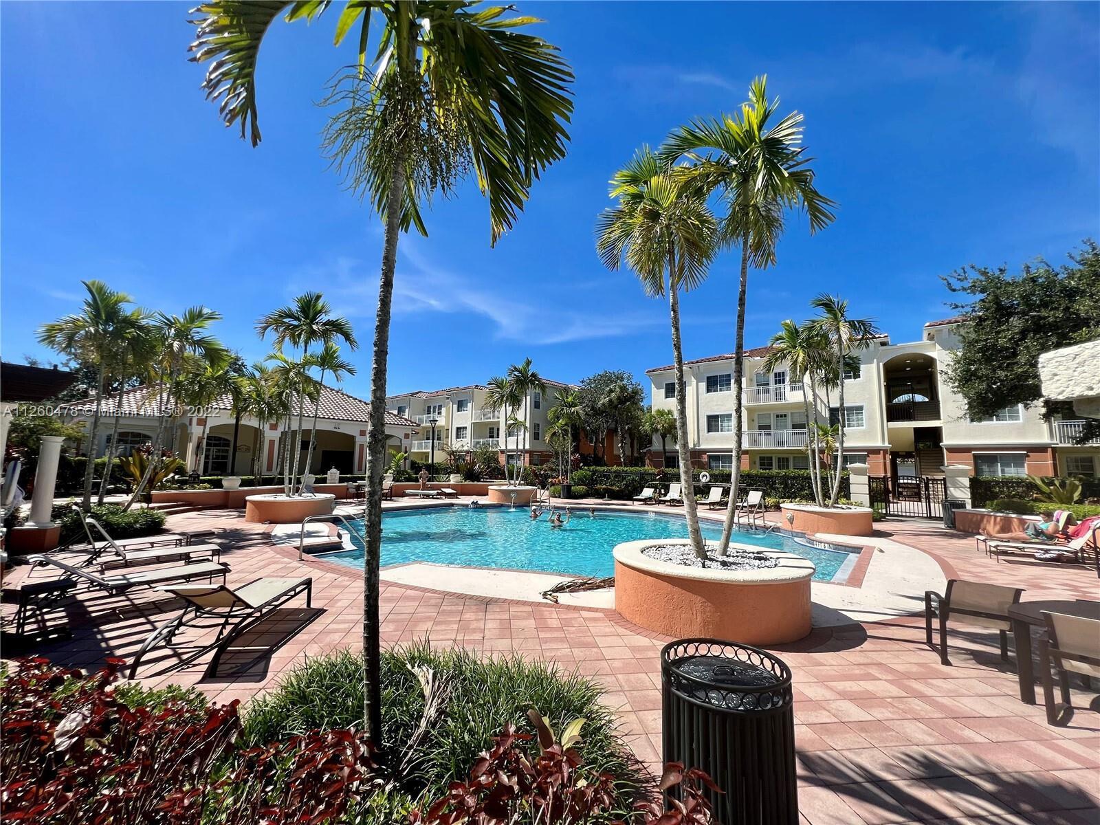 An outdoor swimming pool area surrounded by palm trees, lounge chairs, and apartment buildings — 9887 Baywinds Drive # 4305, West Palm Beach FL 33411, WEST PALM BEACH, FL