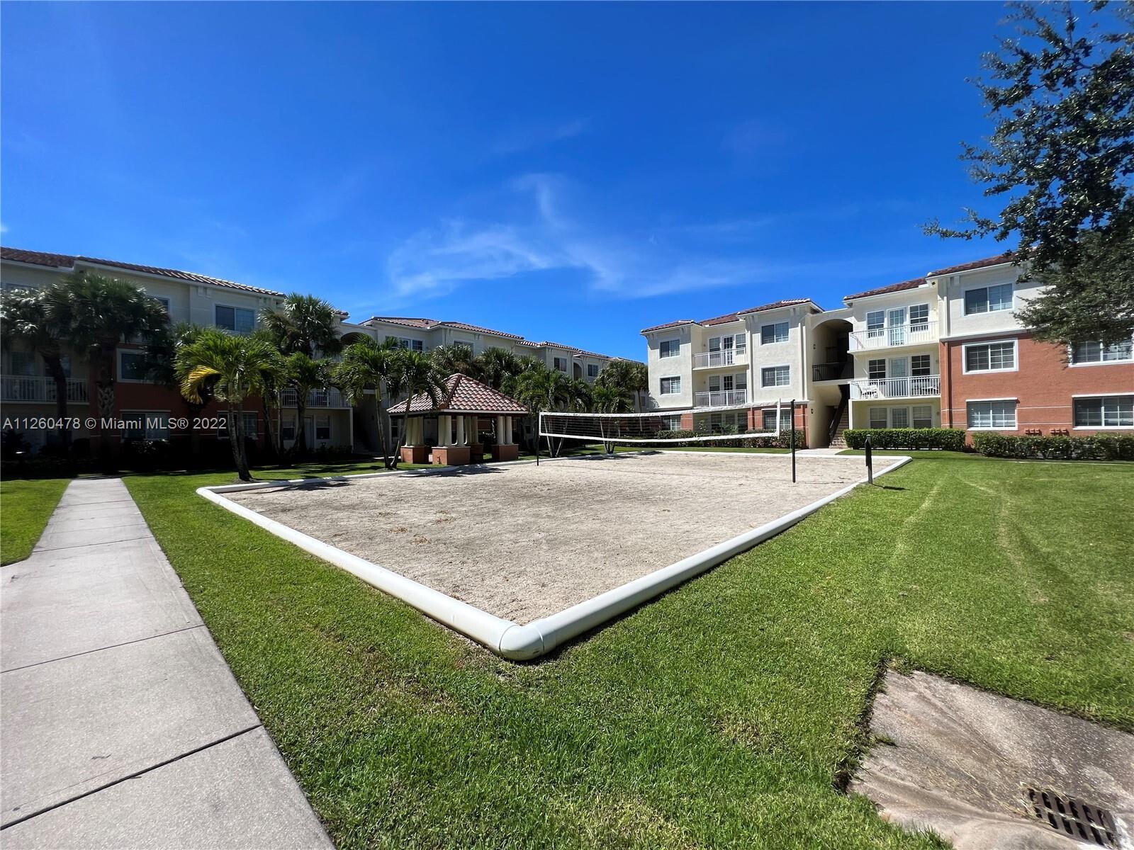A sand volleyball court sits between apartment buildings under a clear blue sky — 9887 Baywinds Drive # 4305, West Palm Beach FL 33411, WEST PALM BEACH, FL