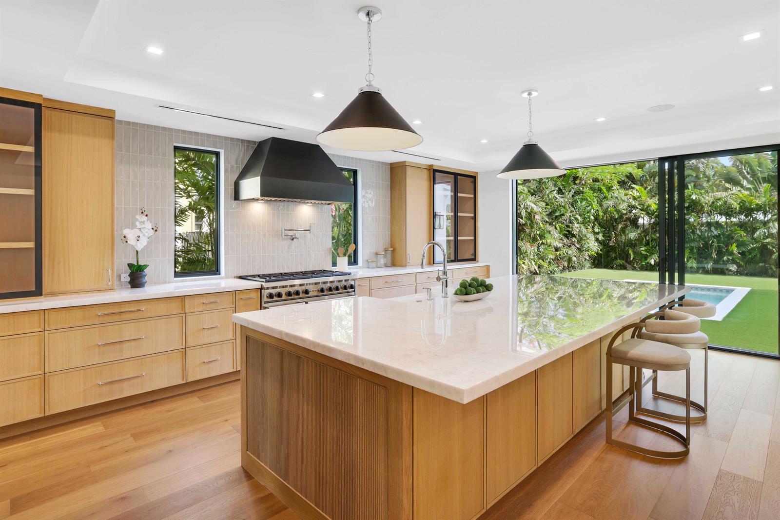 A modern kitchen features light wood cabinetry, a large marble island, and floor-to-ceiling glass doors — 317 Greymon Drive, West Palm Beach FL 33405, WEST PALM BEACH, FL