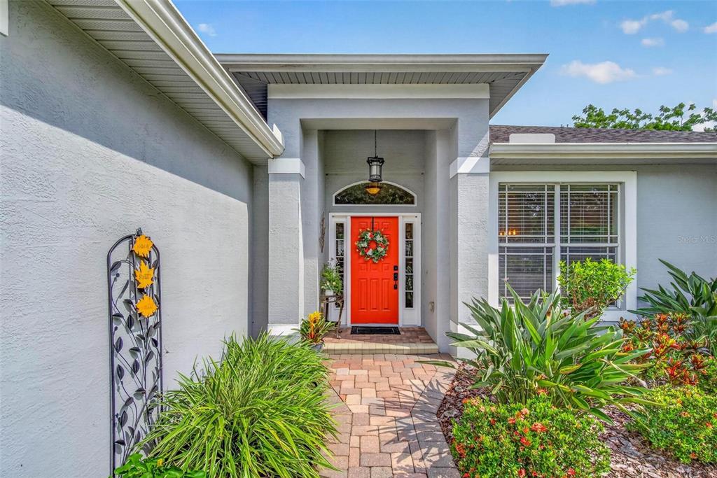 A bright orange front door is centered under a covered entryway with a stone walkway — 2245 MISTY COURT, DUNEDIN, FL