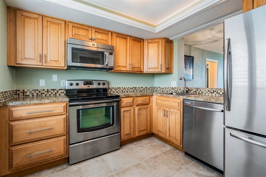 A kitchen features light wood cabinets, stainless steel appliances, granite countertops, and a tiled backsplash — 2620 COVE CAY DRIVE UNIT 601, CLEARWATER, FL