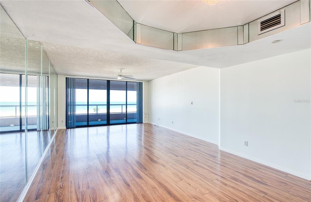 An empty living room features wood-look flooring, mirrored walls, and a sliding glass door to a balcony — 1310 GULF BOULEVARD UNIT 4D, CLEARWATER BEACH, FL