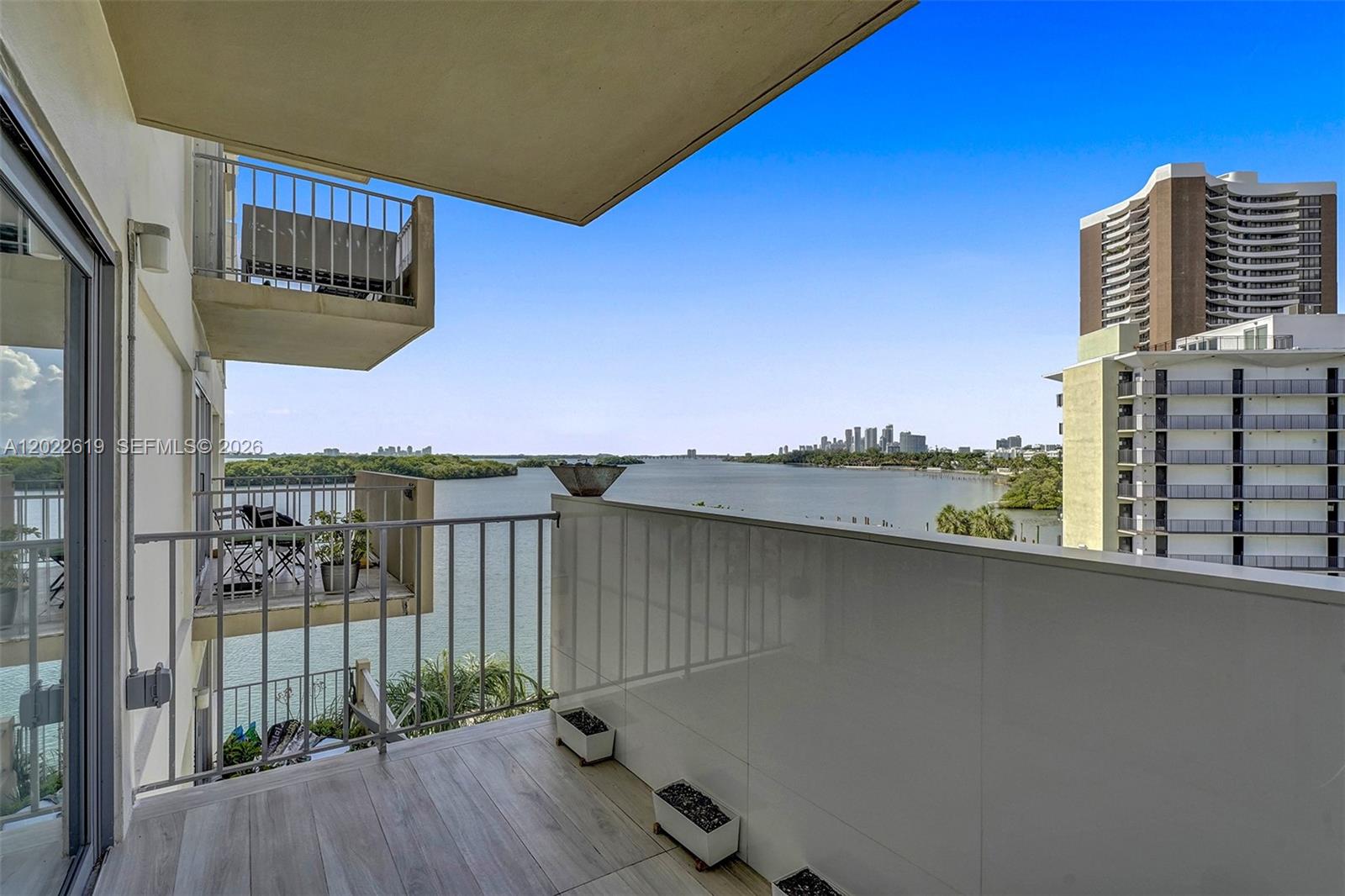 A balcony with wood-look tile flooring overlooks a bay with a distant city skyline — 880 NE 69th St # 5G, Miami FL 33138, MIAMI, FL