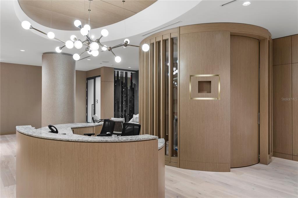 A modern reception area features a curved wooden desk, a chandelier, and light wood flooring — 691 S GULFVIEW BOULEVARD UNIT 1011, CLEARWATER BEACH, FL