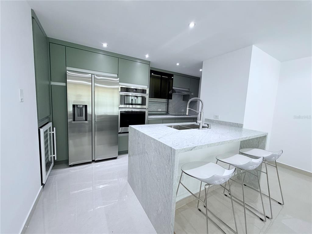 A modern kitchen features sage green cabinetry, stainless steel appliances, and a marble waterfall island — PARKSIDE CII UNIT 906, GUAYNABO, PR