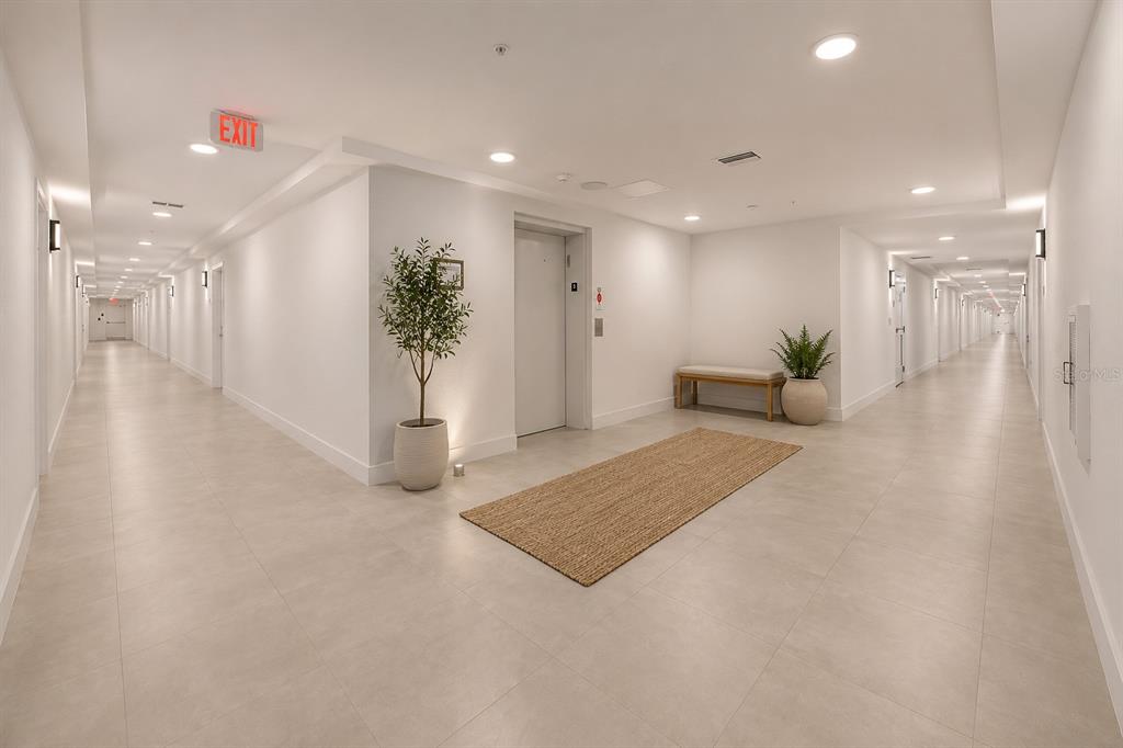 A bright, modern hallway features light tile flooring, white walls, an elevator, and decorative plants — 5587 MILLENIA PARK BOULEVARD UNIT 503, ORLANDO, FL
