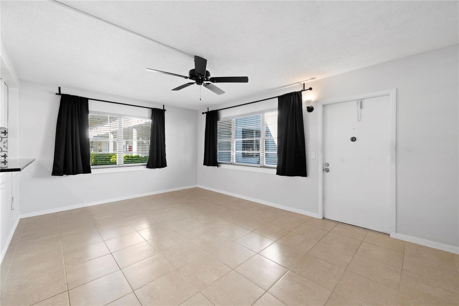An empty living room features white walls, beige tile flooring, black curtains, and a ceiling fan — 28 Prescott B # 28, Deerfield Beach FL 33442, DEERFIELD BEACH, FL