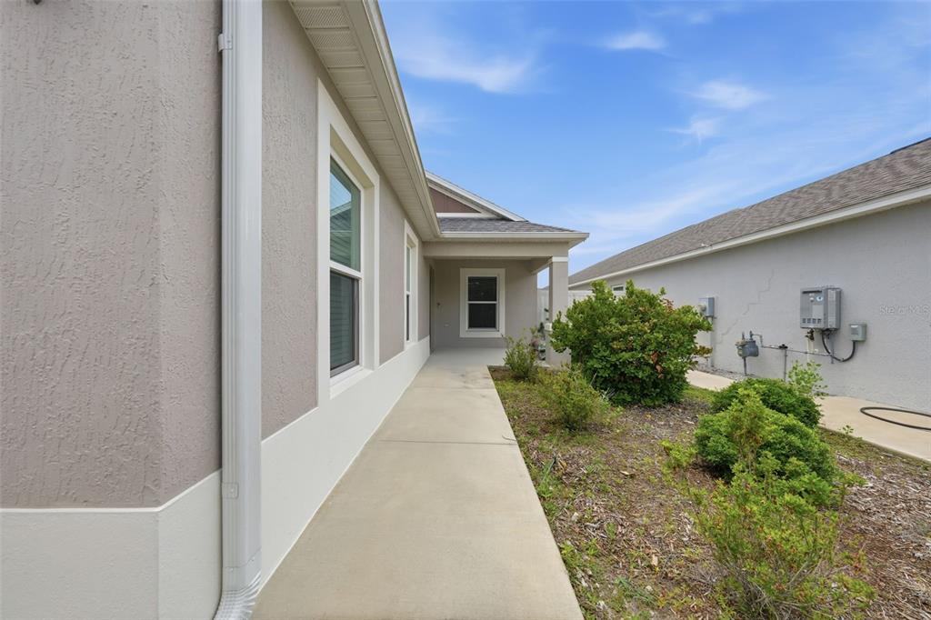 A concrete walkway runs alongside a beige house exterior with a covered entryway and landscaping — 6094 RODRIGUEZ LANE, THE VILLAGES, FL