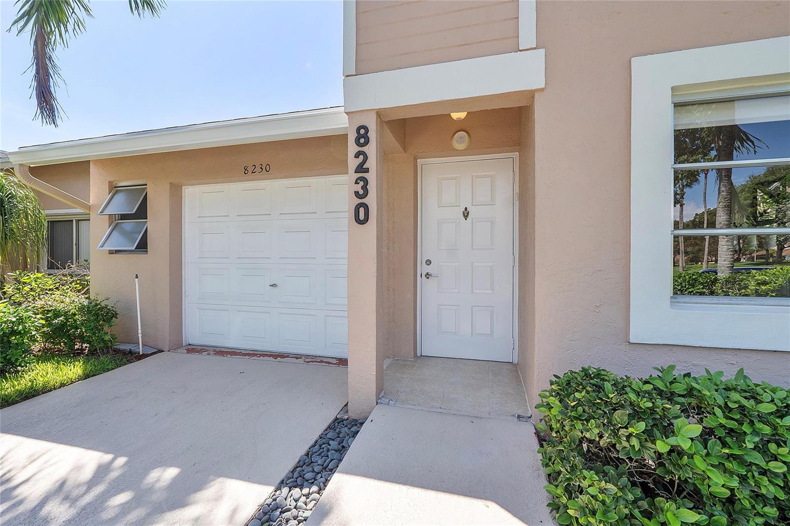 A tan house exterior features a white garage door, a front door, and a window — 8230 Springtree Road, Boca Raton FL 33496, BOCA RATON, FL