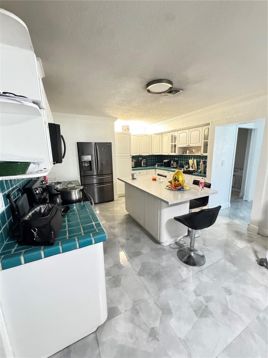 A kitchen features white cabinetry, a central island with bar stools, and teal tiled countertops — 1314 W 77th St, Hialeah FL 33014, HIALEAH, FL