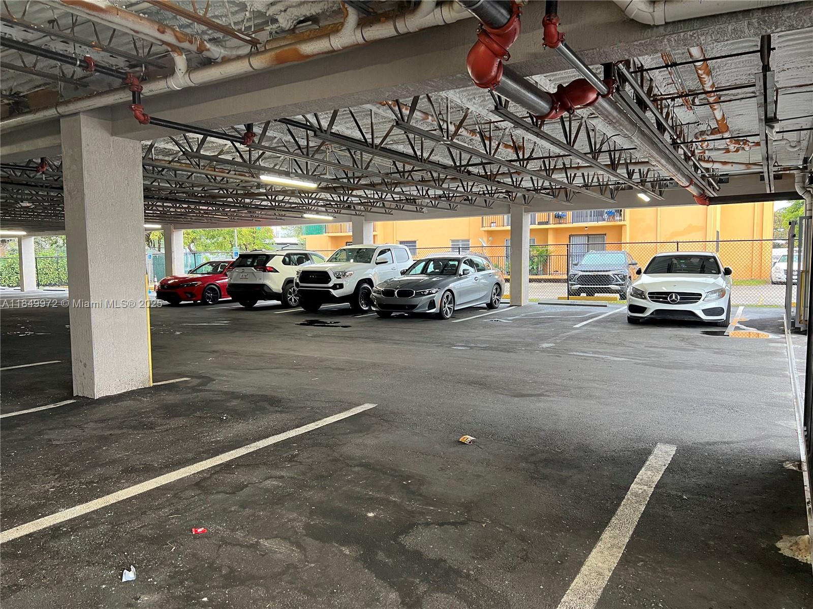 A covered parking garage features several parked cars and exposed overhead pipes and structural beams — 2311 NW 22nd Ave # 207, Miami FL 33142, MIAMI, FL